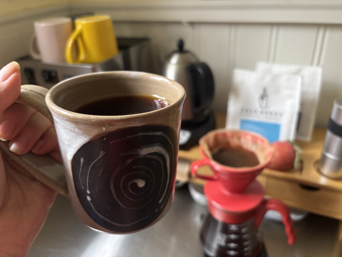 A hand holding a handmade ceramic mug of black coffee, with a red Hario V60 pourover and a bag of beans on the counter behind it.